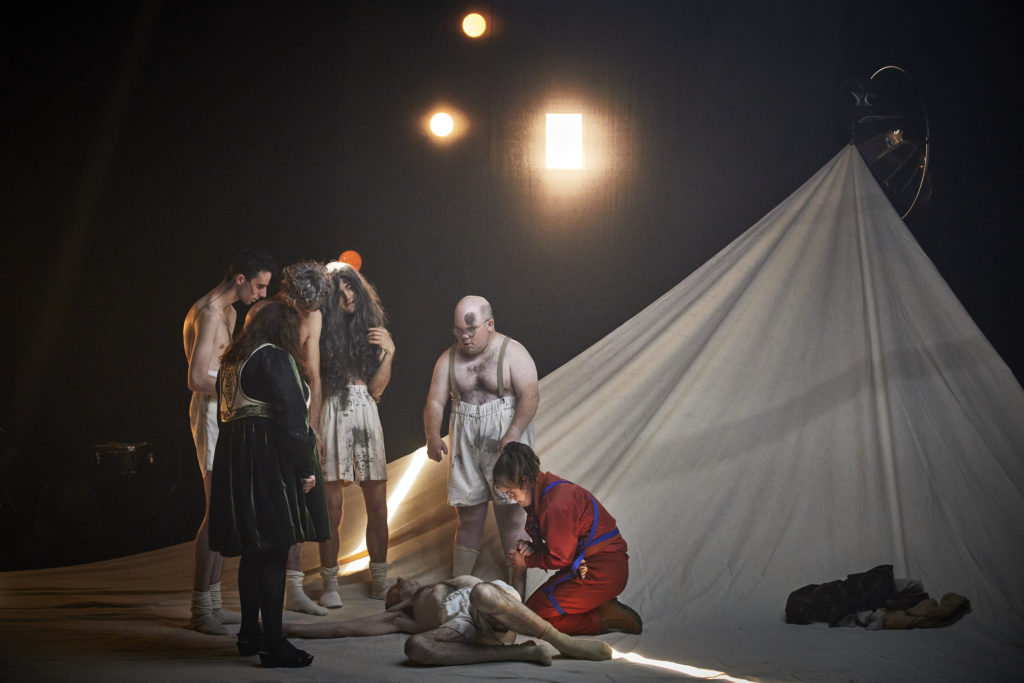 A group of learning disabled actors in tattered costumes stand around another actor injured on the floor