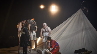 A group of learning disabled actors in tattered costumes stand around another actor injured on the floor