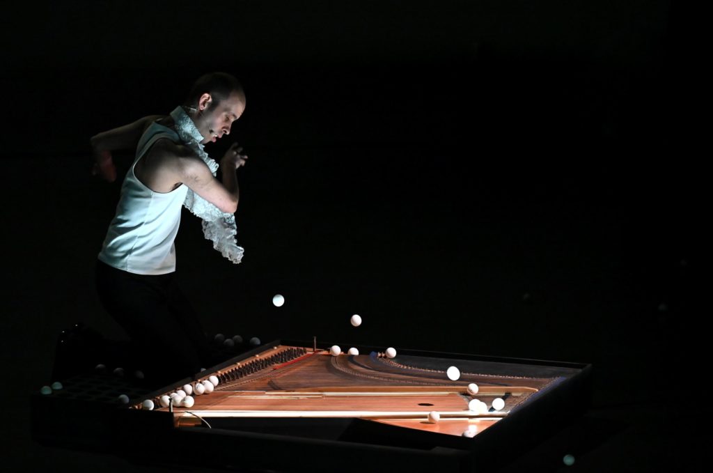 A man stands throwing ping pong balls into a grand piano