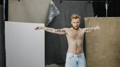 A male performer with one arm has 'I am an artist' written across his body.