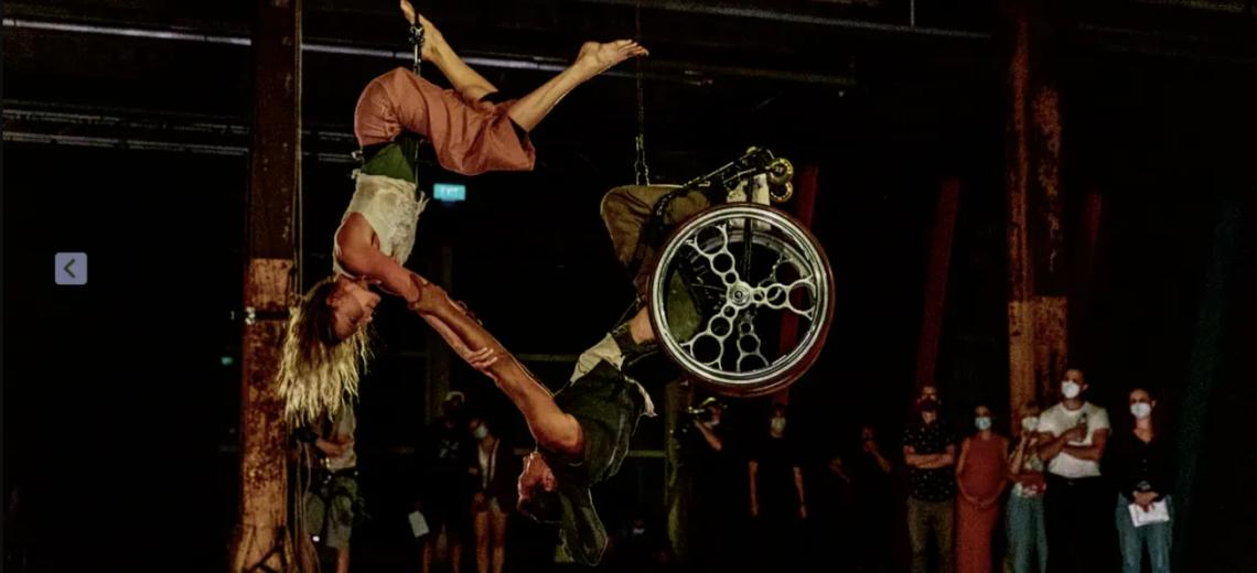 Aerial performance featuring two performers upside down. A Maori male wheelchair user dancer and a white female dancer.