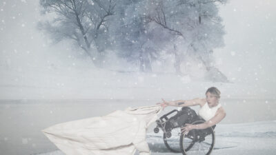A female performer in a wheelchair leans back into a snowy blustery scene.