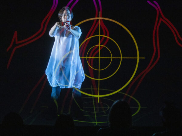 Chisato is on stage wearing a white billowy dress. There is a projection of a yellow target on the black background behind her, along with another red and purple rounded abstract shape. Her two index fingers form a cross, outstretched in front of her.