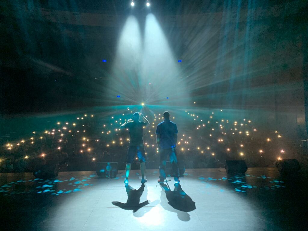 Two performers stand on stage in front of a sea of lights
