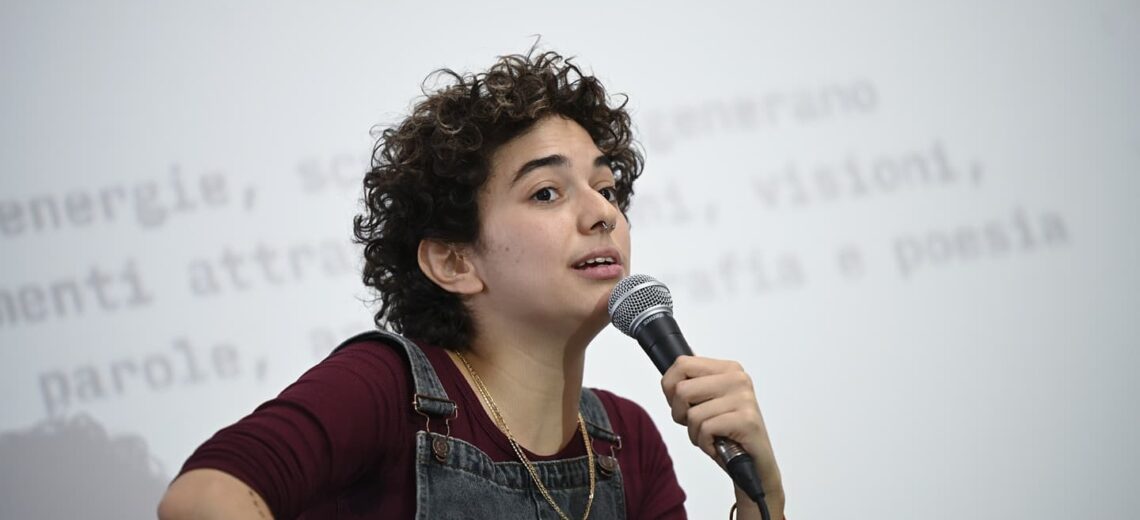 A white non-binary performer with short curly hair speaks into a microphone.