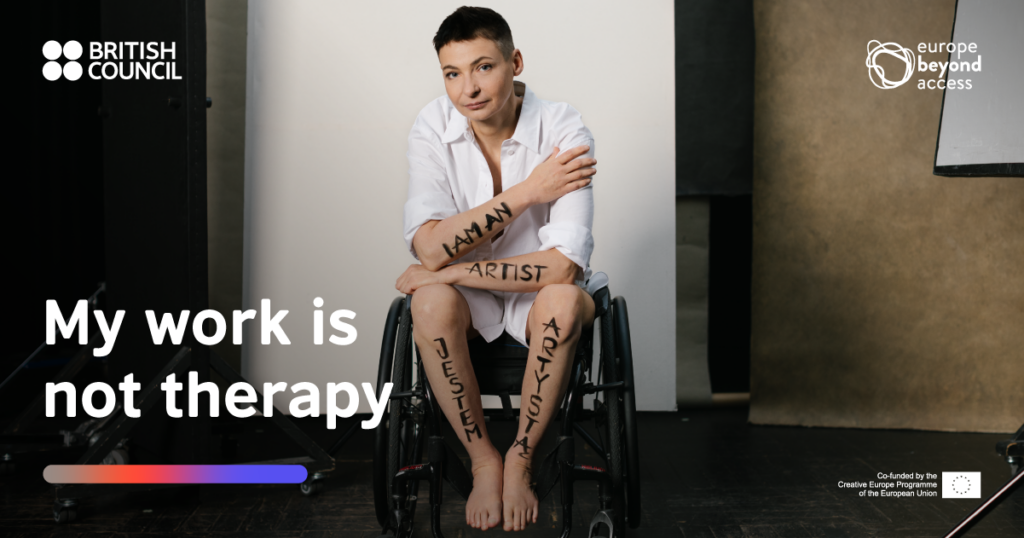 A non-binary performer in a wheelchair sits in a white shirt and the words 'I am an artist' scrawled in black paint on their arms.