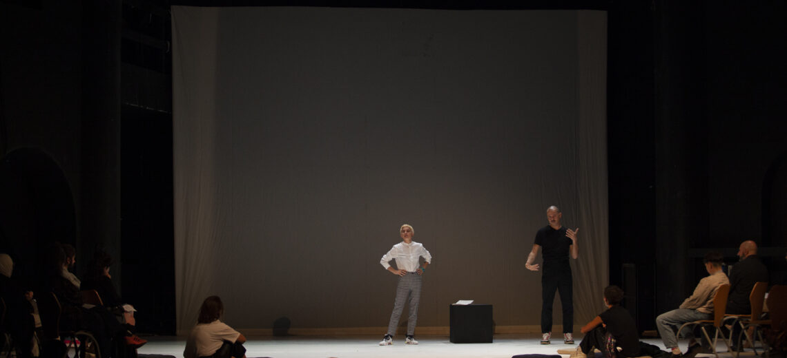 A far out shot of a white non-binary performer stands in a white top and grey trousers on stage.