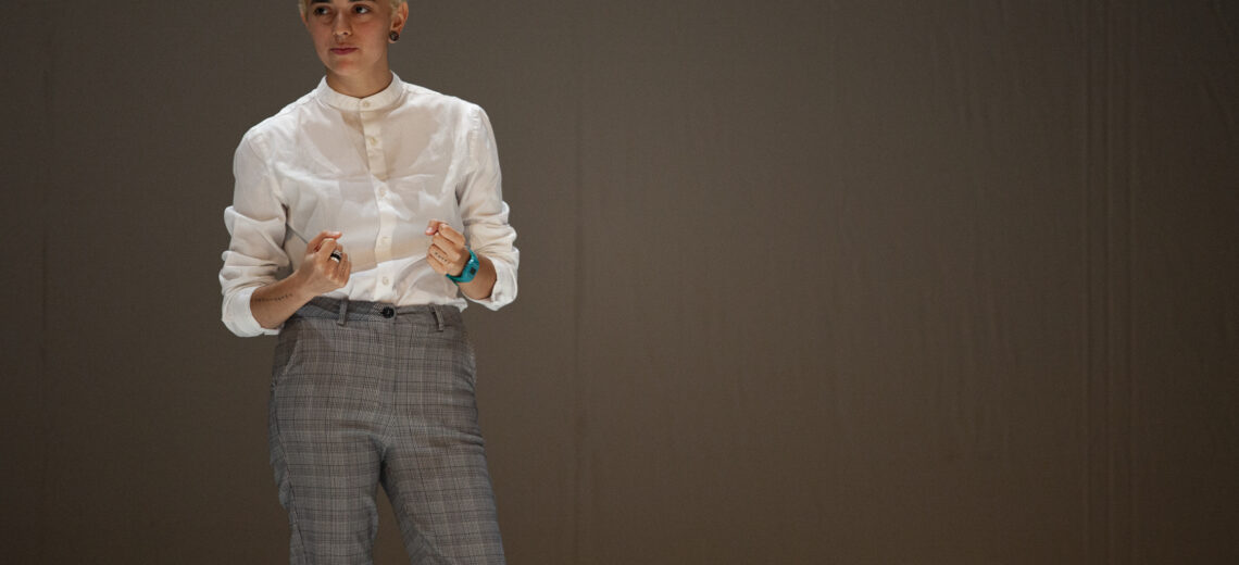 A white non-binary performer stands in a white top and grey trousers on stage.