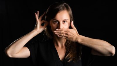 A white female Deaf performer looks direct into the camera, one hand covers her mouth, the other makes a small circle near her ear.