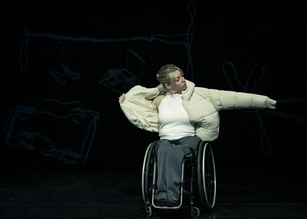 A white female dancer with blonde hair and a wheelchair puts on a cosy padded white winter coat on stage.