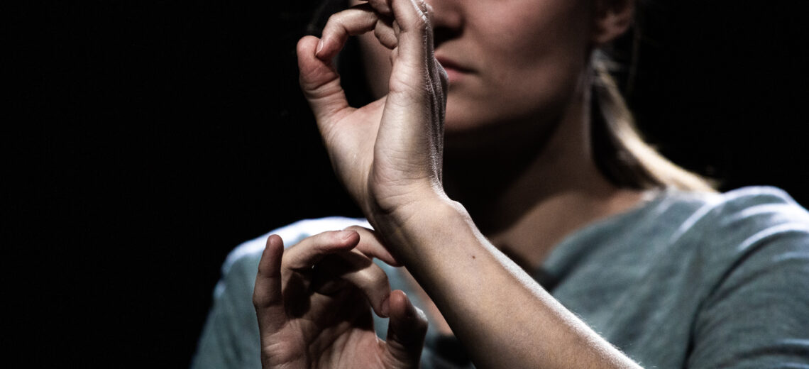 A white Deaf female performer speaks with her hands closely in front of her face