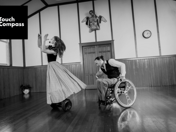 A dance featuring a European female dancer with pale skin and long flowing hair stands upright on a segway, moving right to left. Behind her and in an expressive pose is a Maori male wheelchair user dancer. The poses of the two dancers create physical levels as they dance following each other. From a Touch Compass dance work named Drift featuring Julie van Renen and Rodney Bell. 