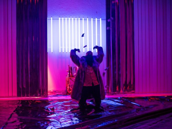 A white male performer lit in pink and purple light, curls his arms above his head