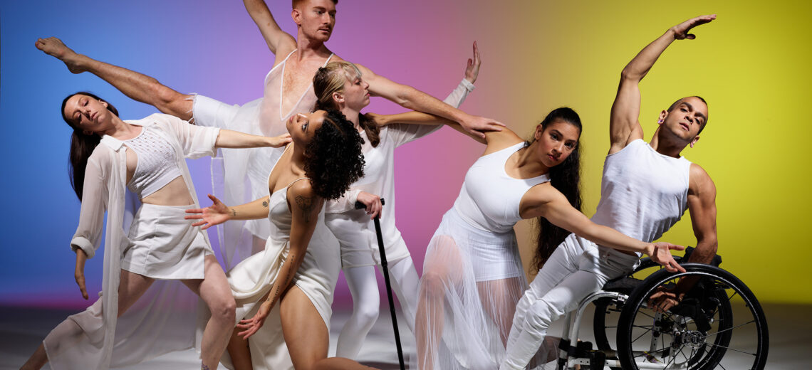 Six disabled and non-disabled dancers pose in white clothes against a rainbow gradient backdrop. The dancers reach, balance and extend various limbs in different directions as they connect to each other.