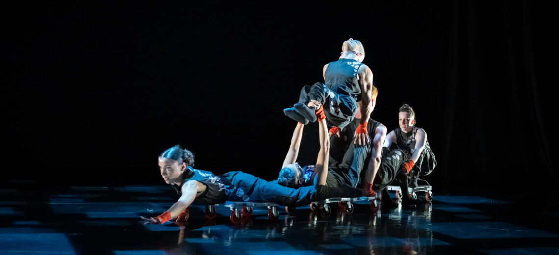 A photo from 'Tread', choreographed by Hewlett 50 collaborating artist Ben Levine. In the photo, David Calhoun sits on a four-wheeled scooterboard, flying through space while JanpiStar lies on their back. Alaja pushes the two dancers forward. The dancers are illuminated by a light pattern made up of various sized squares on a black marley floor.