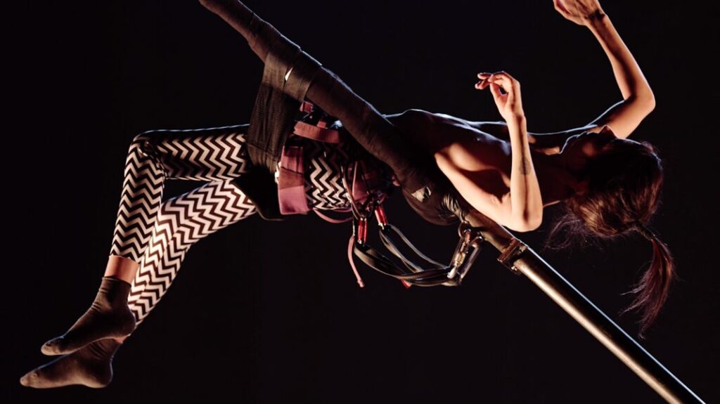 A white petite woman with zigzag tights lies back suspended in the air with poles supporting her.
