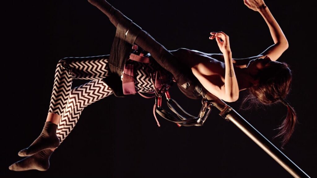 A white woman with zigzag tights leans back suspended in the air and supported by poles.