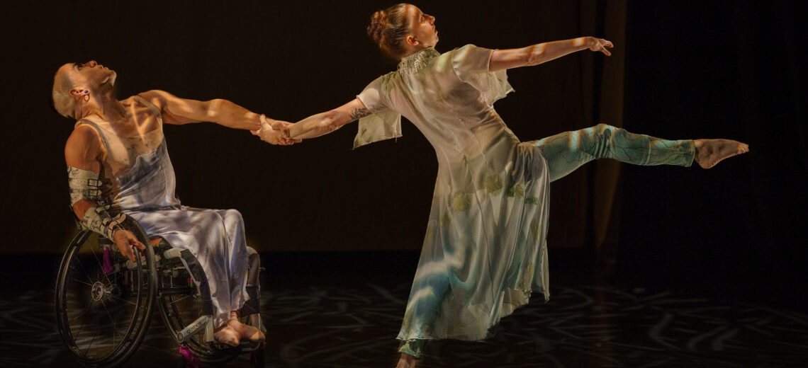Janpi and Louisa hold a counterbalance with hands intertwined as they lean away from each other. Janpi sits in their wheelchair and looks upwards while Louisa reaches one arm and one leg up in attitude. Janpi wears a light purple skirt and tank top, Louisa wears a white and turquoise dress and leggings, illumistaed by dappled stage lighting.