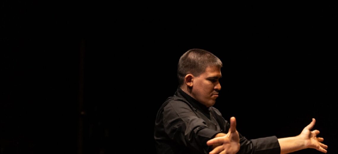 A white disabled male performer conducts an unseen performer with his hands
