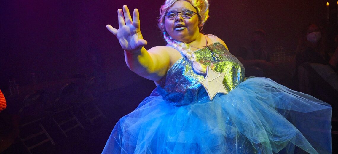 A disabled performer in an over the top blue ball gown sticks out her hand forcefully