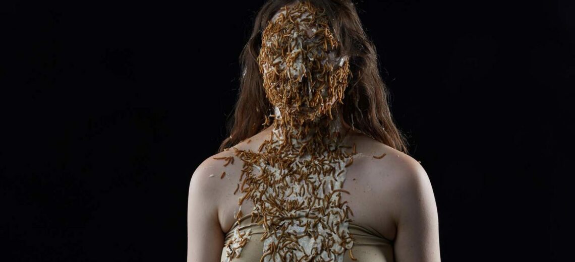 A white female performer, masked, with her face completely concealed with what could be centipedes or fallen plants