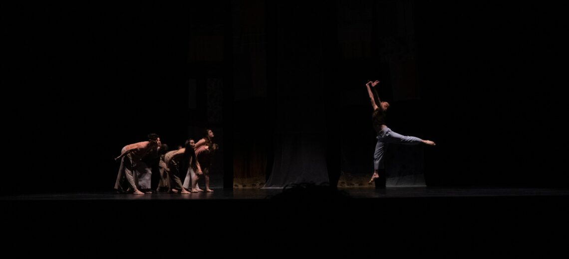 The photo shows a group of dancers doing a circular movement with their backs and another dancer at the other end of the stage doing a jump.  In the background there are some fabrics that make up the scenery of the stage.