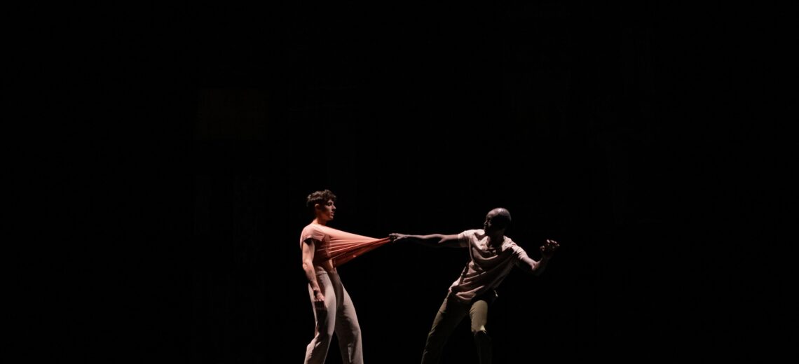 The photograph shows a dark background with two dancers in the center of the scene. One of them tugs at the other's shirt in a sense of opposition.