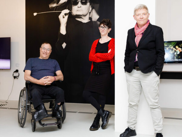White man in a wheelchair and two white women stand in front of a large photograph of someone smoking
