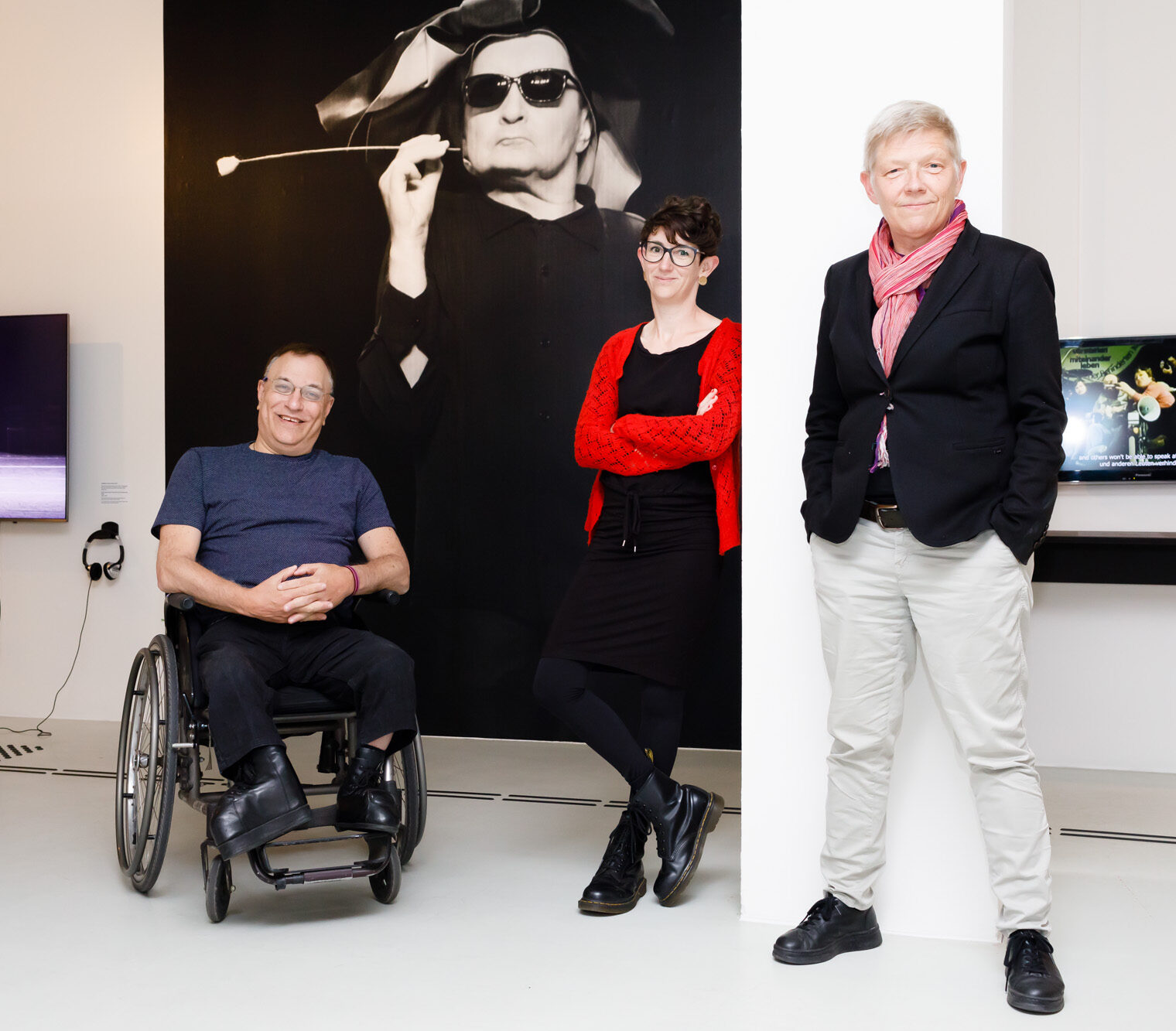 White man in a wheelchair and two white women stand in front of a large photograph of someone smoking 