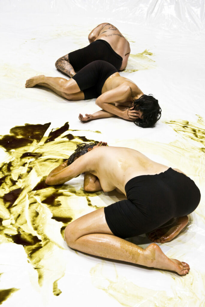 Three performers lie on the floor, covered in brown oily substance, their faces hidden.