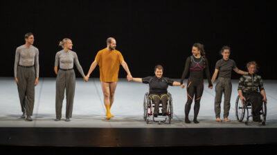 Six performers, including a wheelchair user, two men wearing frocks and three women in trousers and braces perform rapturously to an onlooking crowd in an outdoor space.