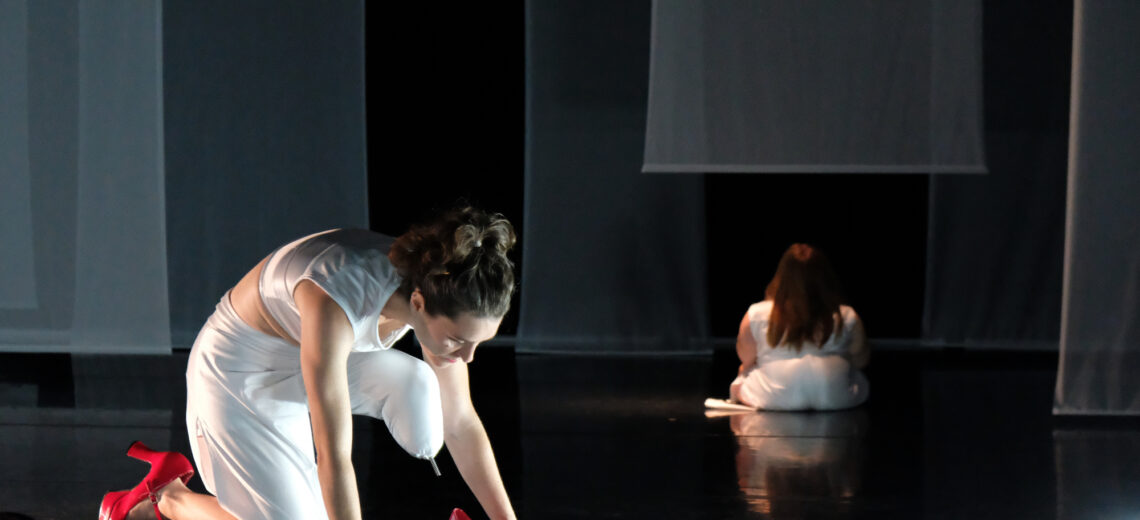 Two female dancers on stage, white dressed. One in front with a half leg prosthesis trying to put on a red shoe and one in the back sitting on the flor with the back to the audience.