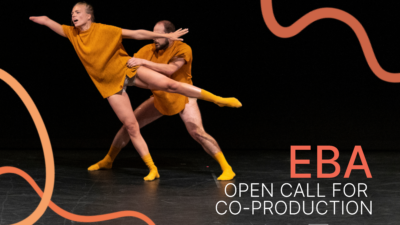 A male dancer holds a female dancer by the waist as she stretches away from him. The female dancer has one arm shorter than the other, and she reaches away in an off-balance position.
