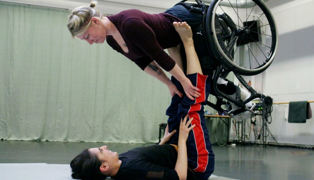 In a dance studio, a female dancer is on her back and lifts a wheelchair female dancer on her feets. They wear gym clothes