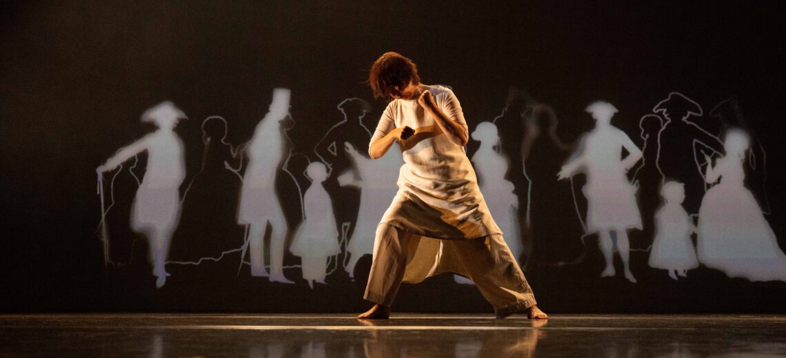 Chisato stands on stage, pitched over as if pulling something heavy on ropes behind her. On the screen behind her are animated images of outlines of figures from 18th Century Europe.