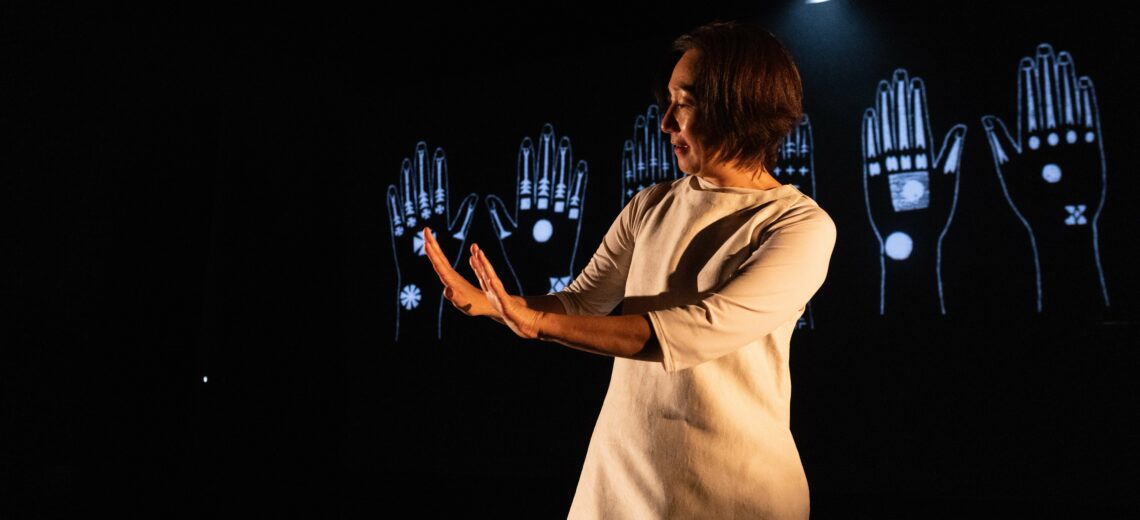 Chisato stands on stage, her hands to side. She is looking at the backs of her hands, as if admiring them. Projected on the screen behind her are animated images of hands with tattoos on them.