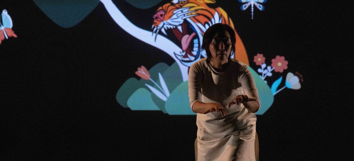 Chisato stands, poised with arms infant of her as if ready to pounce. On the screen behind her us a colourful projection of a roaring tiger surrounded by plants.