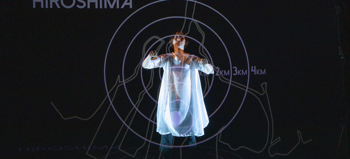 Chisato stands arms outstretched, as if driving a large vehicle. Projected around her are circular lines forming a target over body, and the word 'Hiroshima' is displayed in the upper left corner.