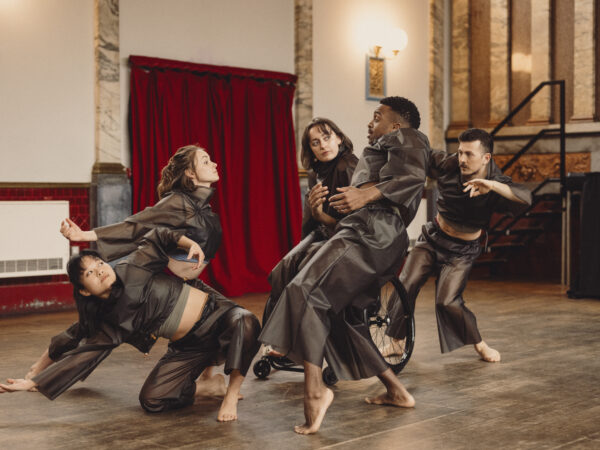 5 dancers moving together, reaching, bending, sitting, lunging in black costumes.